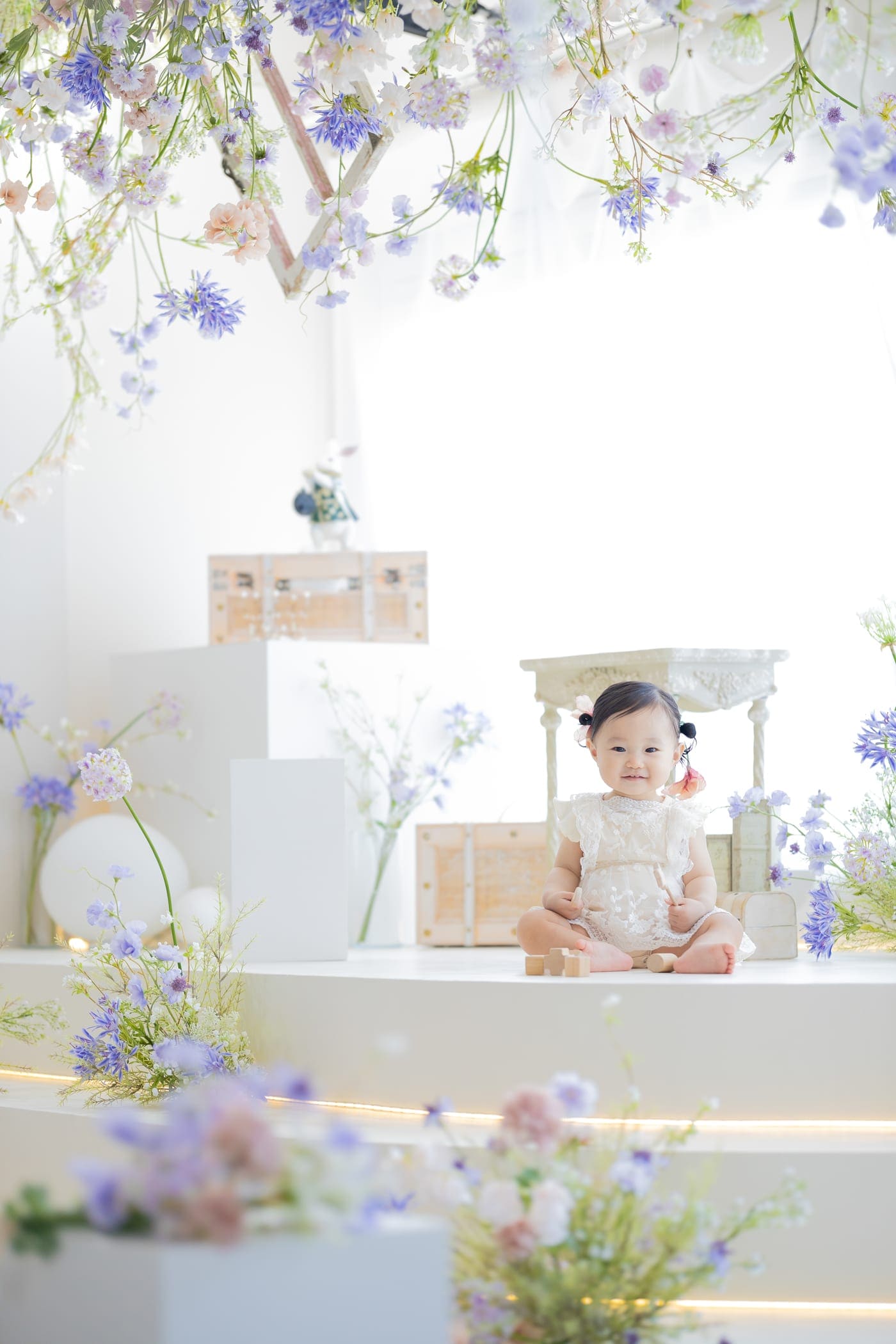 淡い青や紫の花に囲まれた白いスタジオで、レースの衣装を着て座る女の子