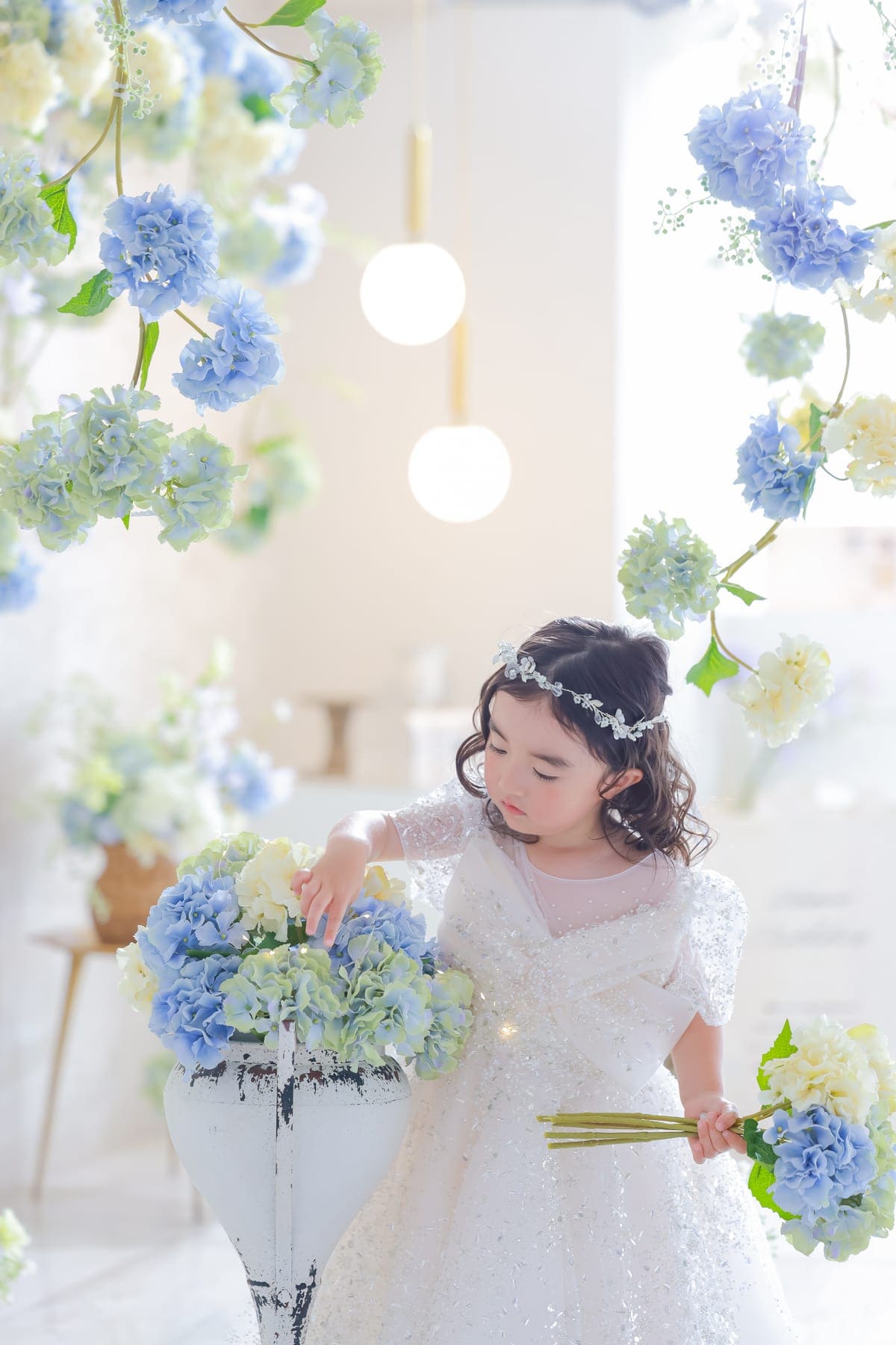 青や白のあじさいに囲まれたスタジオで、白いドレスを着て花に手をのばす女の子