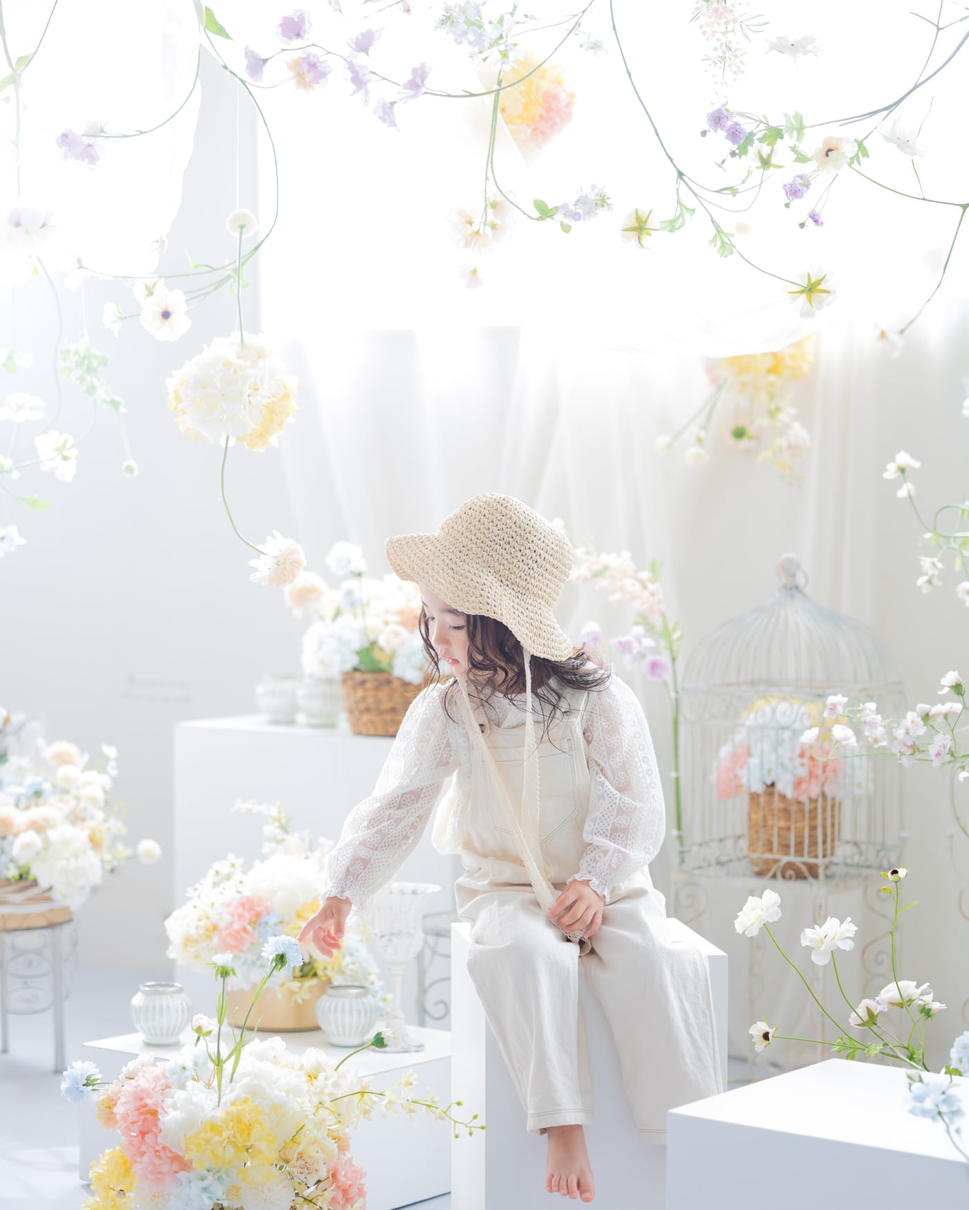 淡い色の花に囲まれた白い空間で、麦わら帽子をかぶって座る女の子