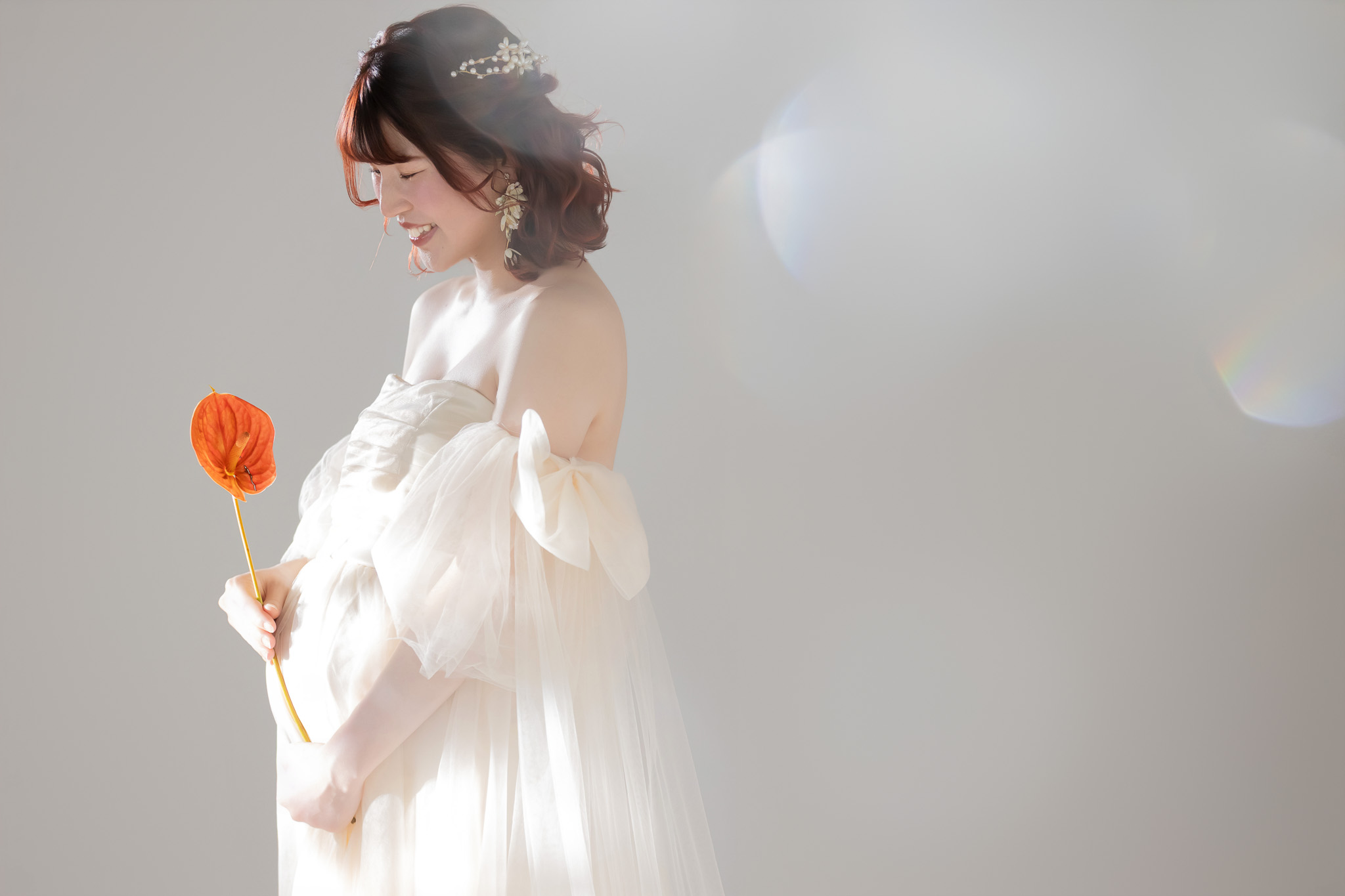 白いチュールドレスをまとい、オレンジの花を手にお腹へ視線を落として微笑む妊婦