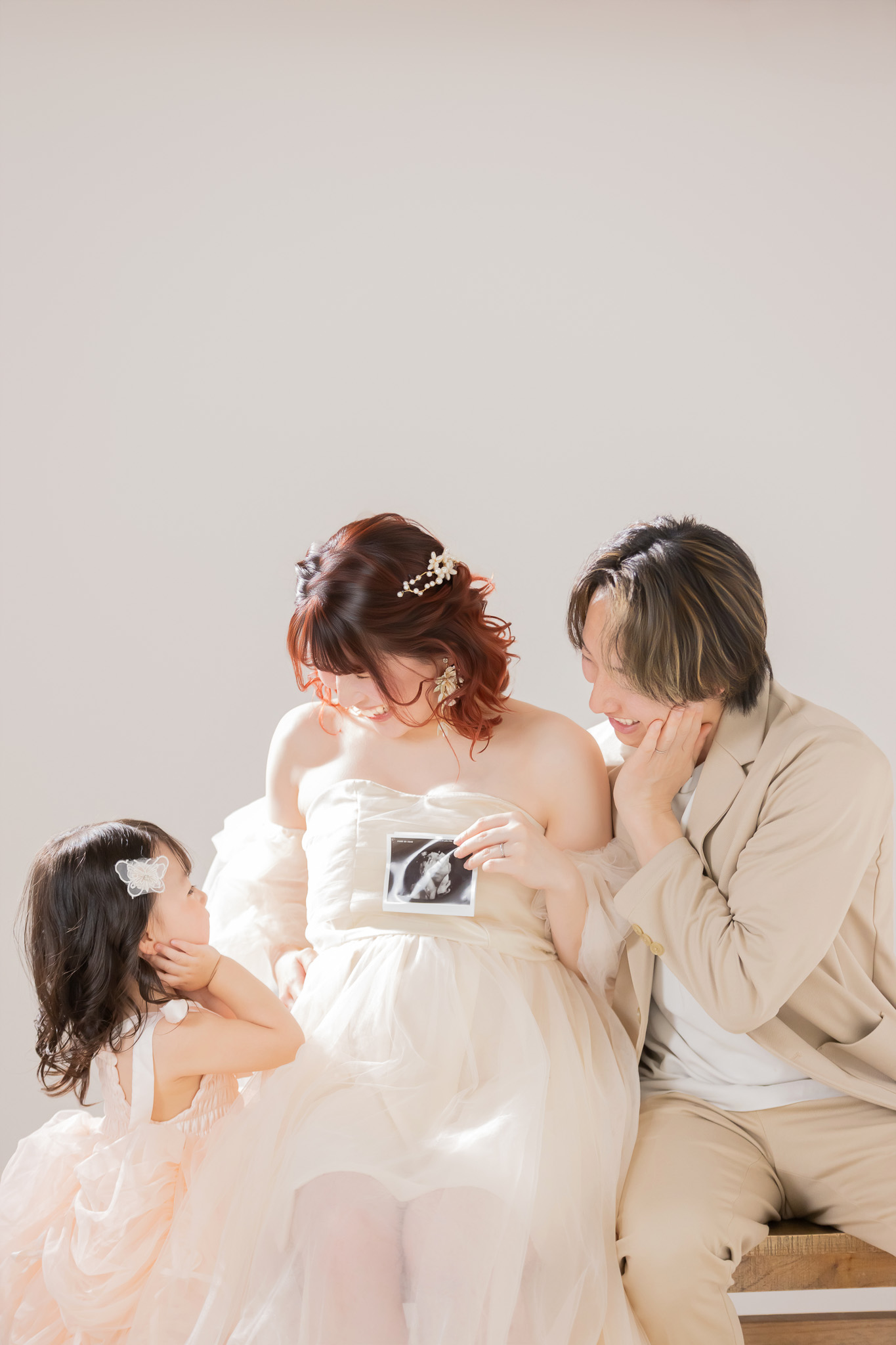 淡いドレスの妊婦がエコー写真を持ち、女の子とベージュのジャケットの夫が両側から寄り添う