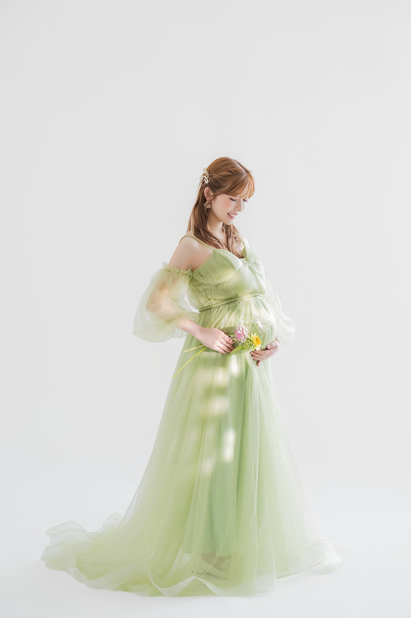 淡いグリーンのチュールドレスで立ち、小さな花束を手にお腹へ視線を落とす妊婦の全身
